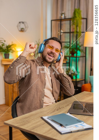 Happy Caucasian man in headphones and sunglasses dancing listening to music at home office desk 128356685