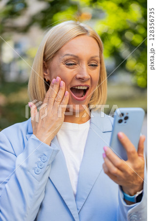 Mature Caucasian businesswoman CEO celebrating good news on smartphone standing on city street Mature Caucasian businesswoman CEO celebrating good news on smartphone standing on city street 128356735