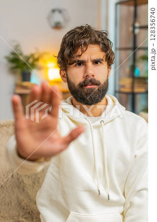 Indian young man guy on sofa couch with serious expression doing stop gesture with hand at home Indian young man guy on sofa couch with serious expression doing stop gesture with hand at home 128356740