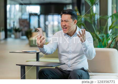 Angry young man boss in shirt holding smartphone arguing over video call in modern office lobby Angry young man boss in shirt holding smartphone arguing over video call in modern office lobby 128356771