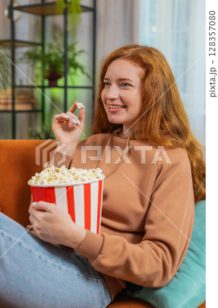 Smiling redhead woman eating popcorn and watching movie wow expression sitting on sofa at home 128357080