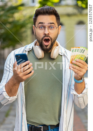 Happy Caucasian man counting money euro cash using smartphone calculator app on city street Happy Caucasian man counting money euro cash using smartphone calculator app on city street 128357087