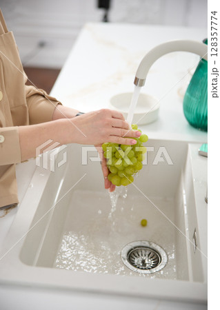 Close up view of a woman washes fresh green grapes under the pressure of flowing water in the white kitchen Close up view of a woman washes fresh green grapes under the pressure of flowing water in the white kitchen 128357774