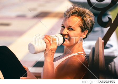 Middle-aged sporty woman sitting on bench and drinking water after cycling in summer sunlight Middle-aged sporty woman sitting on bench and drinking water after cycling in summer sunlight 128358453