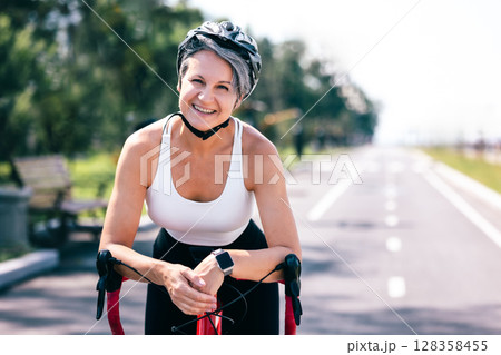 Smiling senior woman on red bicycle in helmet enjoying summer ride on empty park bike lane Smiling senior woman on red bicycle in helmet enjoying summer ride on empty park bike lane 128358455