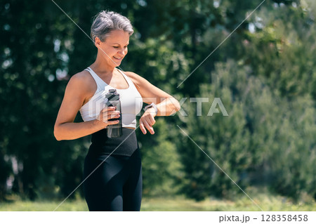 Smiling Mature Woman Checking Fitness Watch While Holding Water Bottle After Outdoor Workout 128358458