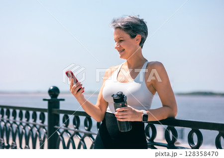 Smiling Active Woman Using Smartphone Outdoors by River with Water Bottle in Hand 128358470
