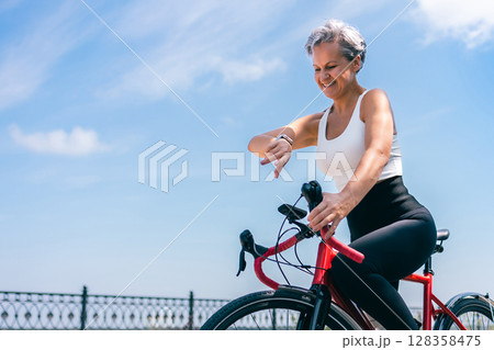 Smiling Senior Woman Checking Smartwatch While Cycling Along Seafront on Sunny Day Smiling Senior Woman Checking Smartwatch While Cycling Along Seafront on Sunny Day 128358475
