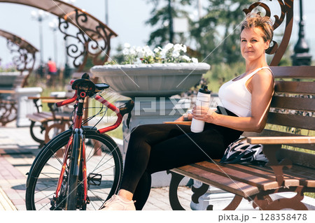 Fit woman resting on bench with bicycle and water bottle during summer ride in urban park Fit woman resting on bench with bicycle and water bottle during summer ride in urban park 128358478