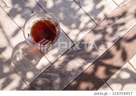 【日本】テラスの木漏れ日が落ちる木製テーブル上に置かれたガラスコップに淹れられたブラックコーヒー 【日本】テラスの木漏れ日が落ちる木製テーブル上に置かれたガラスコップに淹れられたブラックコーヒー 128358957