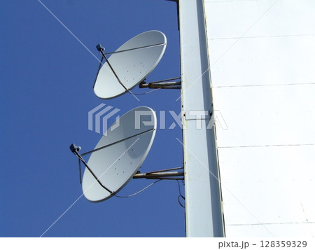 Two white satellite dishes are affixed to the side of a tall structure, facing the clear sky. They are positioned to capture and transmit signals from satellites. 128359329