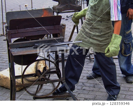 A skilled artisan works at a traditional blacksmith workshop, shaping metal with tools while the forge glows brightly. The setting is filled with handmade creations and tools. 128359346