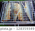 Four fish are placed on a barbecue grill, cooking over an open flame in a sunny outdoor environment. The fish appear golden and crispy, ready for serving. 128359349