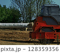 A roller compactor is actively working on a construction site, flattening and compacting soil under a clear blue sky. Nearby, wrapped materials are prepared for use, highlighting ongoing development. 128359356