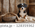 A cheerful puppy with a black and white coat sits next to a large bowl filled with dog food. The background features rustic wooden walls and a warm, cozy atmosphere. 128359362