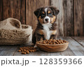 A small, fluffy puppy looks curiously at the camera while sitting beside a wooden bowl filled with pet food. The warm, rustic background enhances the cozy atmosphere of this moment. 128359366