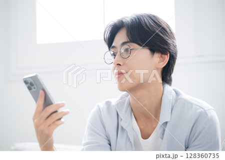 逆光の綺麗な部屋でスマホを見る、持つタップする眼鏡のハンサムな男性のクローズアップ	 128360735