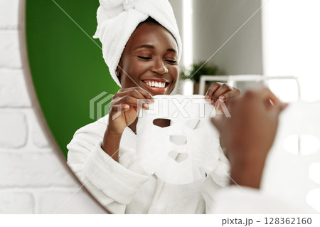 Woman applying a facial mask while smiling in a bathroom setting with a mirror reflection Woman applying a facial mask while smiling in a bathroom setting with a mirror reflection 128362160