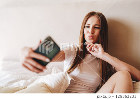 Woman taking a selfie while sitting on bed with white sheets in a cozy indoor setting during daytime Woman taking a selfie while sitting on bed with white sheets in a cozy indoor setting during daytime 128362233