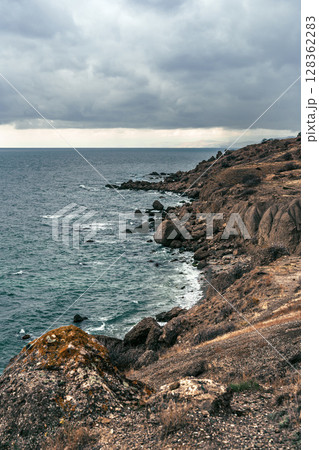 Coastal landscape under cloudy skies with rocky shoreline and gentle waves along the shore Coastal landscape under cloudy skies with rocky shoreline and gentle waves along the shore 128362283