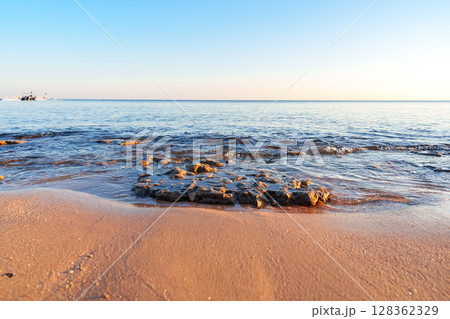 Beautiful sandy shore at sunset with gentle waves lapping against the rocks in a peaceful coastal setting Beautiful sandy shore at sunset with gentle waves lapping against the rocks in a peaceful coastal setting 128362329