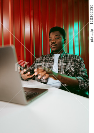 Focused young man working on a laptop while using a smartphone in a vibrant workspace Focused young man working on a laptop while using a smartphone in a vibrant workspace 128362359