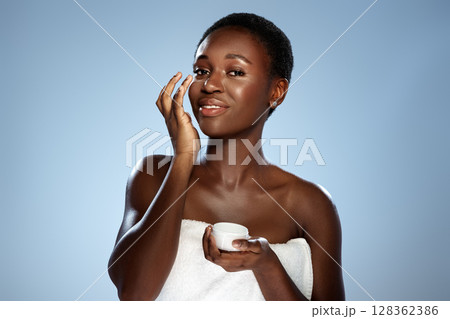Woman applying skincare product while smiling against a light blue background during a beauty routine in a bright indoor setting Woman applying skincare product while smiling against a light blue background during a beauty routine in a bright indoor setting 128362386