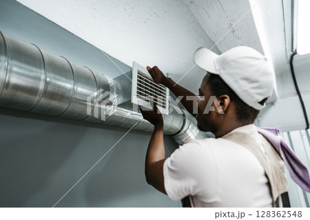 Worker installs ventilation grate in commercial building during day, ensuring proper air circulation for occupants Worker installs ventilation grate in commercial building during day, ensuring proper air circulation for occupants 128362548