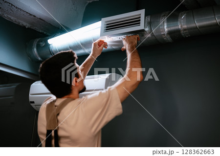 Worker installs air vent in modern indoor space during renovation process in urban environment Worker installs air vent in modern indoor space during renovation process in urban environment 128362663