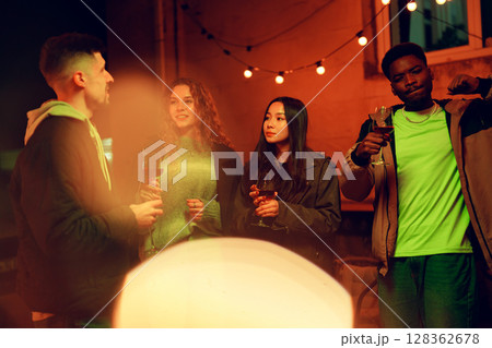 Friends gather around a warm outdoor gathering enjoying drinks and laughter at night under string lights Friends gather around a warm outdoor gathering enjoying drinks and laughter at night under string lights 128362678