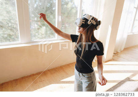 Young woman experiencing virtual reality in a sunlit room with large windows and wooden flooring Young woman experiencing virtual reality in a sunlit room with large windows and wooden flooring 128362733