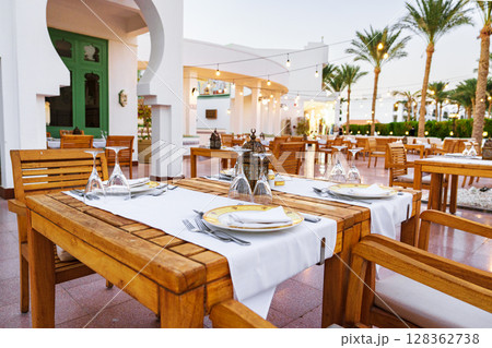 Dining area at a stylish Mediterranean restaurant during sunset with wooden tables and palm trees Dining area at a stylish Mediterranean restaurant during sunset with wooden tables and palm trees 128362738
