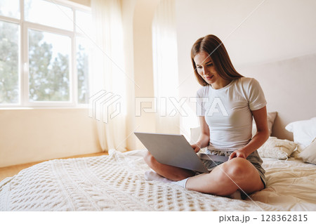 Young woman enjoys online activities while sitting on bed in a bright, airy room during the daytime 128362815