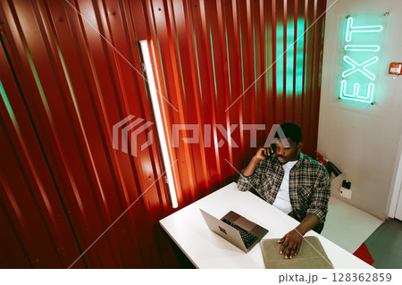 Working professional engaged in a phone call while using a laptop in a modern workspace with distinct wall colors Working professional engaged in a phone call while using a laptop in a modern workspace with distinct wall colors 128362859
