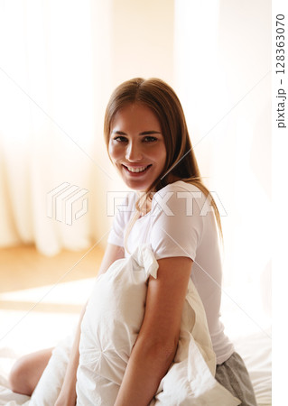 Young woman smiling while holding a pillow in a bright, cozy bedroom during morning light 128363070