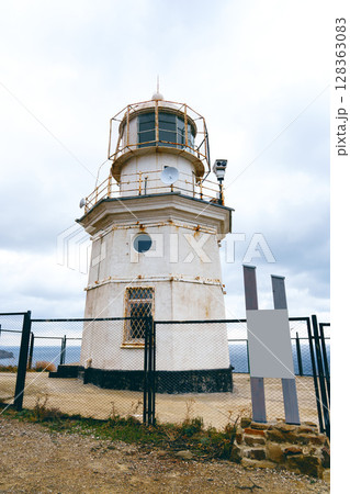 Historic lighthouse stands on rocky coast under cloudy sky with surrounding fence and lookout equipment Historic lighthouse stands on rocky coast under cloudy sky with surrounding fence and lookout equipment 128363083