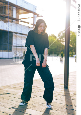 Young woman in casual black outfit poses confidently in urban setting during golden hour 128363098