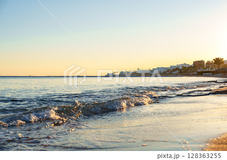 Gentle waves lap against the shore at sunset along a tranquil beach in a coastal town Gentle waves lap against the shore at sunset along a tranquil beach in a coastal town 128363255