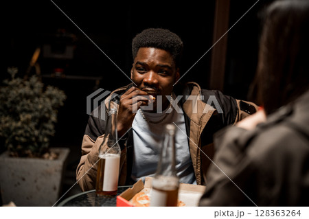Young man enjoying snack outdoors at night with friends while having drinks and conversing 128363264