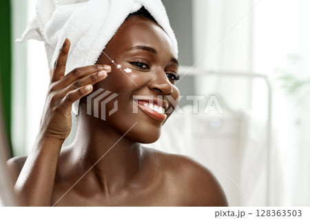 Woman applying skincare products with a towel on her head in a bright bathroom during morning routine 128363503