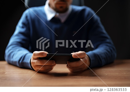 Man focused on smartphone while sitting at a desk in an office setting during the day 128363533
