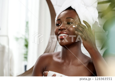 Woman applying moisturizer in a bright bathroom with a towel on her head during skincare routine 128363551