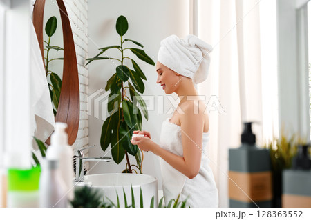 Woman in towel applying skincare product in modern bathroom near window with plants Woman in towel applying skincare product in modern bathroom near window with plants 128363552