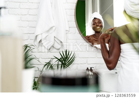 Woman applying skincare in a bright bathroom with modern decor and greenery 128363559