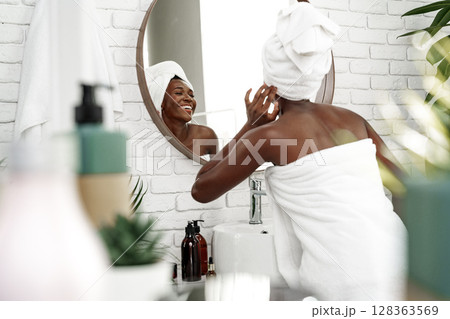 Woman in towel applying skin care in modern bathroom with round mirror and plants at midday 128363569