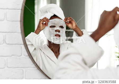 Woman applies a facial mask while looking in the mirror at home during a self-care routine Woman applies a facial mask while looking in the mirror at home during a self-care routine 128363588