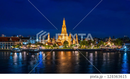 Night view of Wat Arun temple beautifully illuminated on the Chao Phraya River with glowing reflections and city lights in the background 128363615