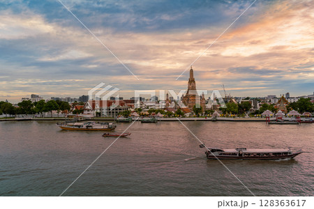 Sunset view of Wat Arun temple across the Chao Phraya River with boats passing by under a dramatic cloudy sky 128363617