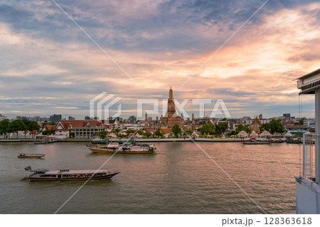 Sunset view of Wat Arun temple across the Chao Phraya River with boats passing by under a dramatic cloudy sky 128363618