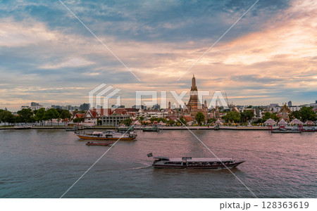 Evening view of Wat Arun temple across the Chao Phraya River with boats passing by under a dramatic sunset sky 128363619